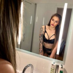 Woman in black lingerie gazes at her reflection in a bathroom mirror, tattoos visible on one arm.